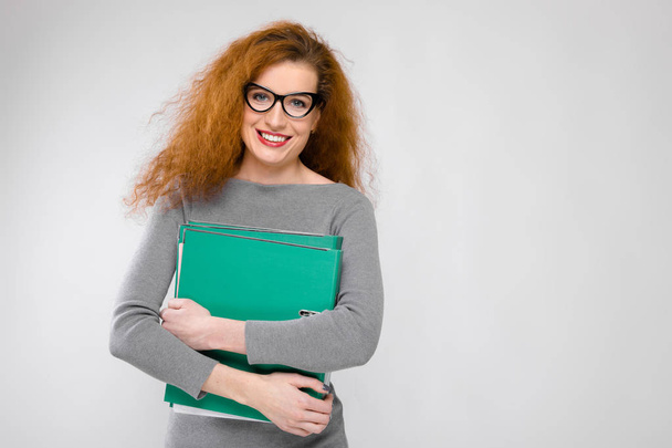 Beautiful curly redhead caucasian woman in glasses and grey dress showing different emotional expressions on white wall in studio. Smiling and holding stack of green folders. - 写真・画像