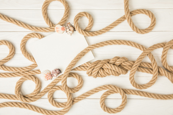 top view of empty paper with seashells on brown nautical knotted ropes on white wooden surface - Foto, imagen
