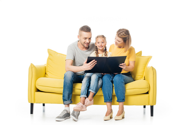 Eltern betrachten Fotos im Fotoalbum, während sie mit Tochter auf gelbem Sofa sitzen - Foto, Bild