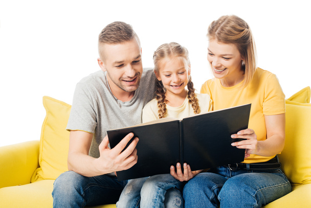 happy family looking at photos in photo album together while sitting on yellow sofa isolated on white - Фото, изображение
