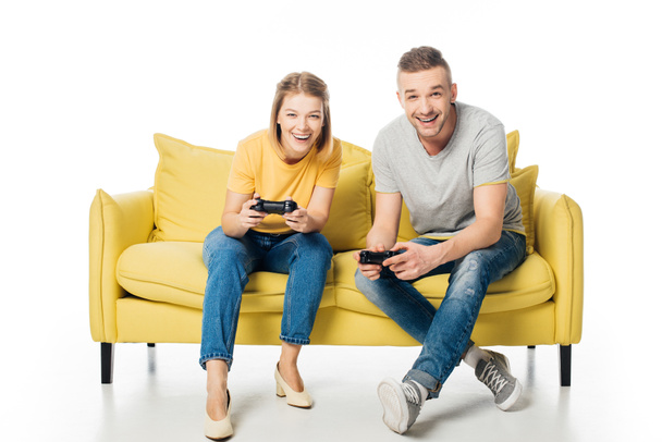 cheerful couple with joysticks playing video game while sitting together on yellow sofa, isolated on white - Φωτογραφία, εικόνα