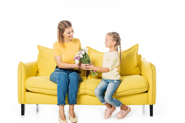 Kleines Kind schenkt Tulpenstrauß an Mutter auf gelbem Sofa isoliert auf weißem, Muttertagsferienkonzept - Foto, Bild