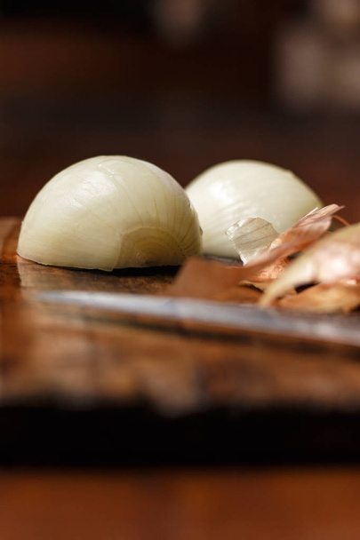 Onion cut in half, onion peels in the foreground and cut knife in the background. Objects on cutting board and rustic wooden table. - Photo, Image