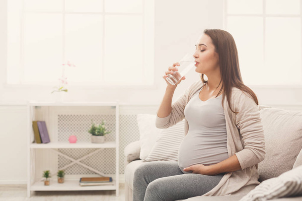 Беременная женщина пьет воду сидя на диване
 - Фото, изображение