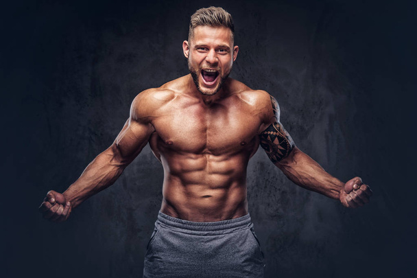 A handsome shirtless bodybuilder with stylish haircut and beard, with tattoo on his arm, shows a war cry in a studio. Isolated on a dark background. - Fotoğraf, Görsel