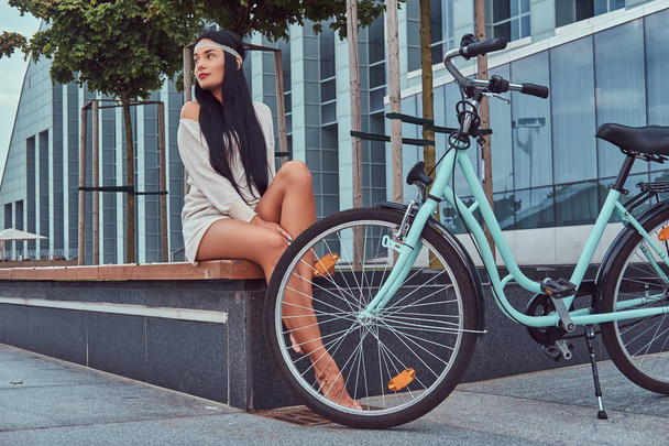 Portrait of a sexy hippie female wearing blouse and shorts in a headband, sits barefoot on a bench near city bike against a skyscraper. - Photo, image
