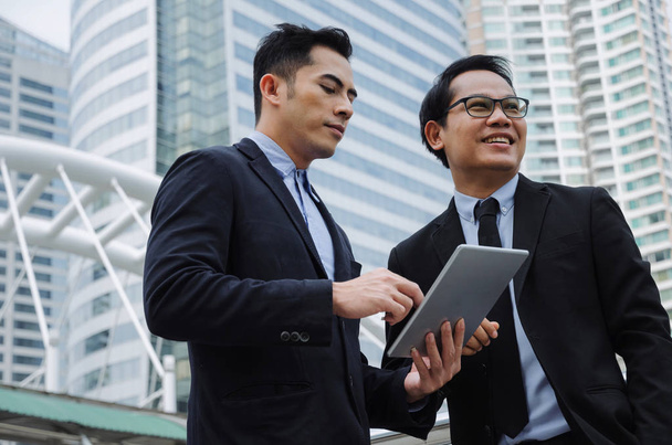 two business young asian smart man talking and reading information about finance news in mobile tablet together standing in modern city, network technology, internet, successful, teamwork concept - Valokuva, kuva