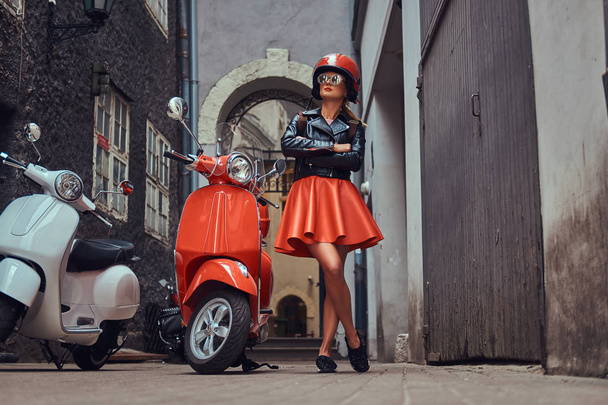 Una chica rubia sexy con ropa elegante en gafas de sol y casco, de pie con los brazos cruzados en una vieja calle estrecha con dos scooters retro
. - Foto, Imagen