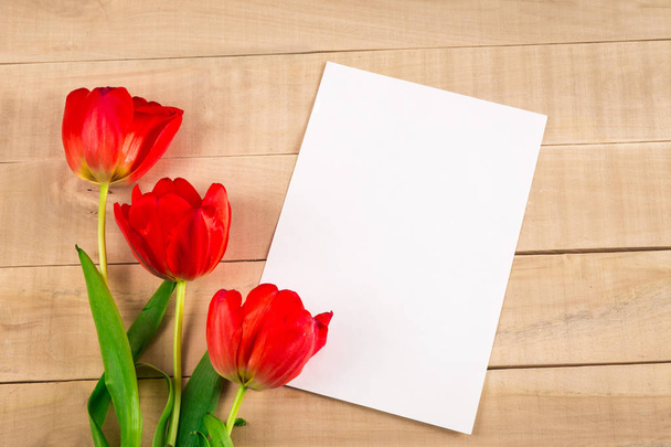White sheet of paper and three red tulips lie on a wooden table - Φωτογραφία, εικόνα