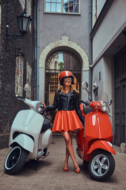 A sexy blonde girl wearing stylish clothes in sunglasses and helmet, standing on an old narrow street with two retro scooters. - Photo, Image