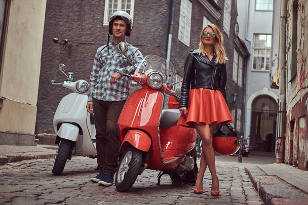 An attractive couple, a handsome man and sexy female standing on an old street with two retro scooters. - Photo, Image