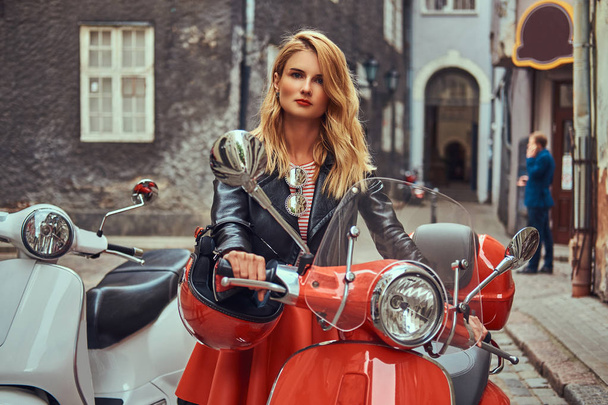 A sexy blonde girl wearing stylish clothes standing on an old street with two retro scooters. - Photo, Image