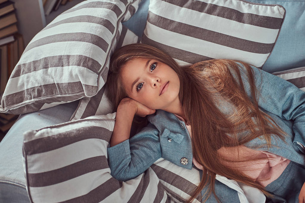 Portrait of a cute little girl with long brown hair and piercing glance, looking at a camera, lying on a sofa at home alone. - Valokuva, kuva