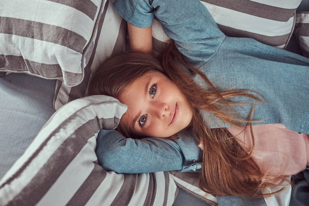 Retrato de una linda niña de cabello castaño largo, mirada penetrante y sonrisa encantadora, mirando a una cámara, tumbada en un sofá en casa sola
. - Foto, Imagen