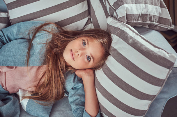 Portret van een schattig klein meisje met lang bruin haar en doordringende blik, op zoek naar een camera, liggend op een bank thuis alleen. - Foto, afbeelding