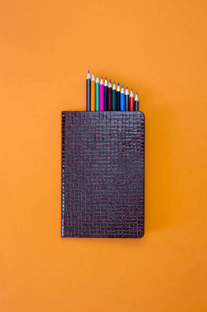 Zárt Jegyzetfüzet-val egy sor színes ceruzák, narancsszínű háttérrel, felülnézet - Fotó, kép