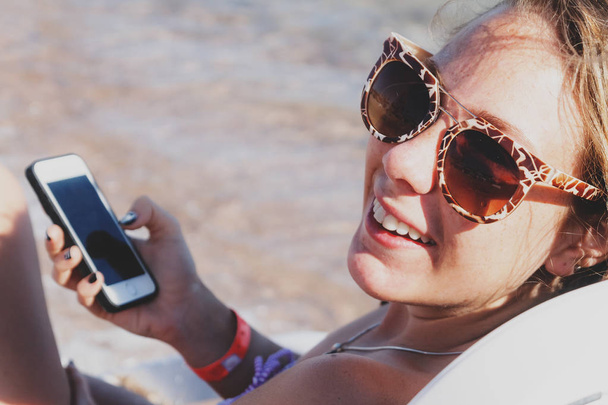 Молодая загорелая женщина смотрит и смеется в смартфон через
  - Фото, изображение