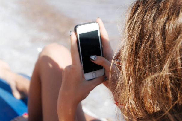 Egy fiatal cserzett nő néz egy smartphone keresztül napszemüveg  - Fotó, kép