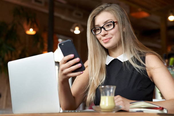 επιτυχημένη επιχειρηματίας νέων γραπτών μηνυμάτων σε ένα smartphone που κάθεται σε ένα καφέ με ένα φορητό υπολογιστή και ένα φλιτζάνι τσάι ή καφέ.  - Φωτογραφία, εικόνα