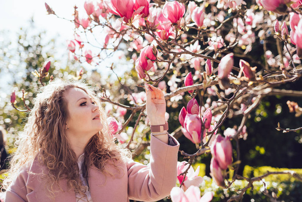 curly young woman walking through the spring park, enjoying the scent of pink magnolia - Zdjęcie, obraz
