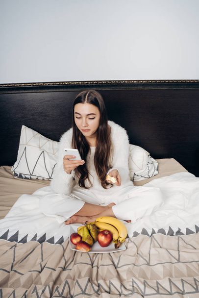 cute long-haired girl in white pajamas eating fruit in bed and looking into her smartphone - Photo, image