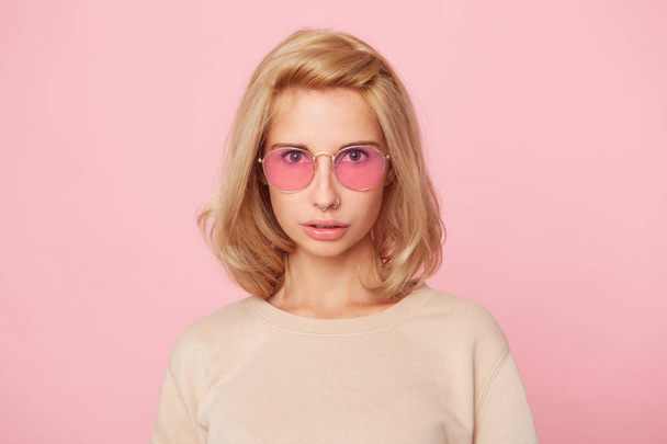 Indoor shot of young blonde female looks directly into camera seriously and angry. Facial expression. Isolated over pink background - Φωτογραφία, εικόνα