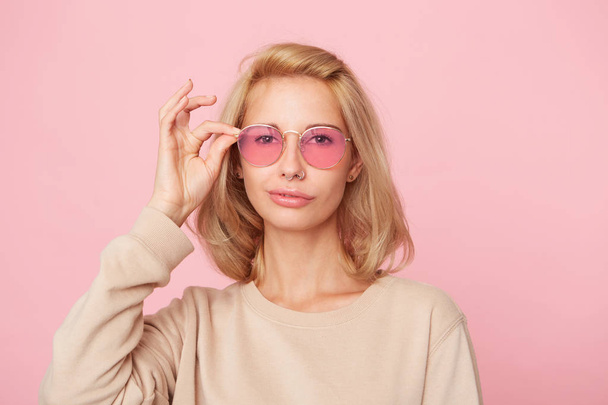 Young blonde woman, shy and smile, stares through spectacles, wears yellow sweatshirt. Isolated over pink background  - Photo, Image