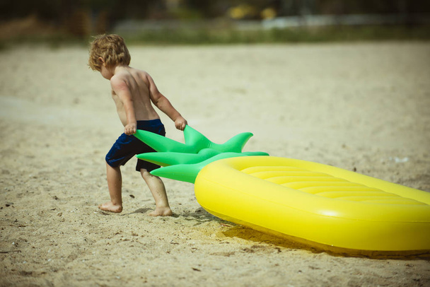 Cute child dragging air mattress at seashore. Toddler on vacation, summer holidays going to swim on mattress. Strong kid dragging pineapple shaped mattress on sand beach. Small and strong concept. - Φωτογραφία, εικόνα