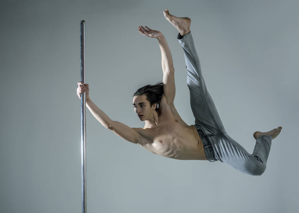 Man with long hair and nude torso wears denim pants, grey background. Athlete, sportsman performing pole dancing moves, work out, show trick. Guy makes figure on pole, fly in air. Work out concept. - 写真・画像
