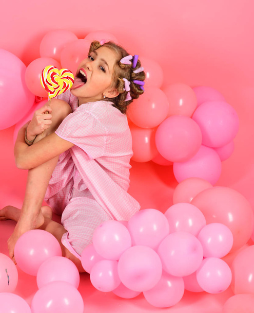 Childhood, happiness, sweet dreams. Small girl child eat lollipop on pink. Little girl with candy lollipop. Diet, birthday, punchy pastels, beauty. Party balloons, kid in curlers, pajama fashion. - Fotografie, Obrázek
