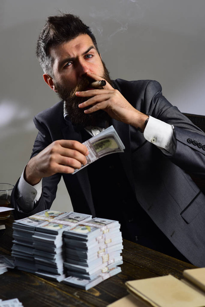 Man sitting at table with piles of money, counting profit. Illegal cash concept. Man in suit smoking cigar while counting money. Businessman on serious face holds money, grey background. - Photo, Image