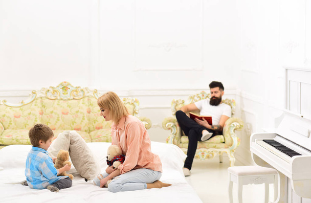 Father not pay attention to kid. Mom and son spend time together in bedroom, luxury interior background. Mother play with cute little son, while father busy with book. Indifferent father concept. - Φωτογραφία, εικόνα