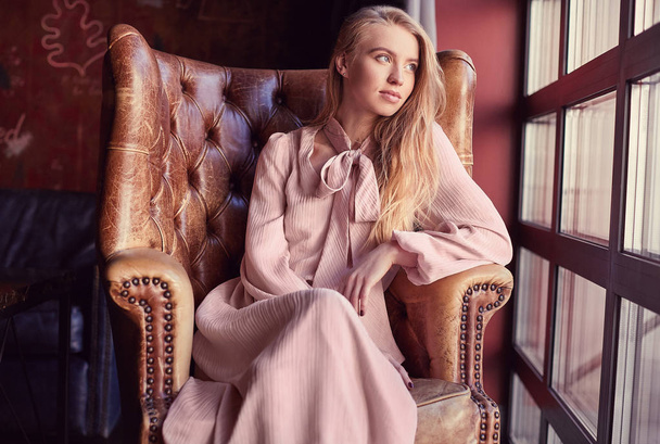 Красивая элегантная блондинка в розовом платье сидит в кожаном кресле
 - Фото, изображение