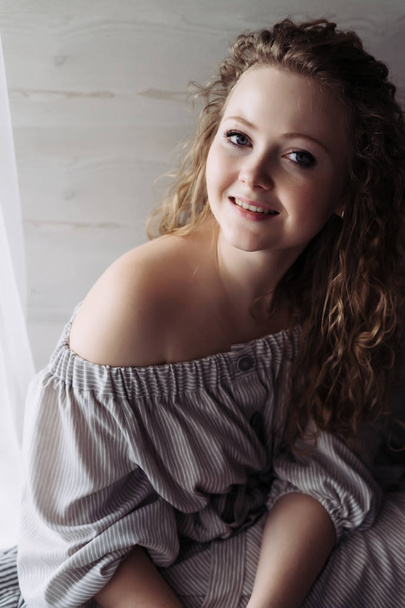 young curly woman in a beautiful dress sits by the window, smiling and looking at the camera - Fotoğraf, Görsel