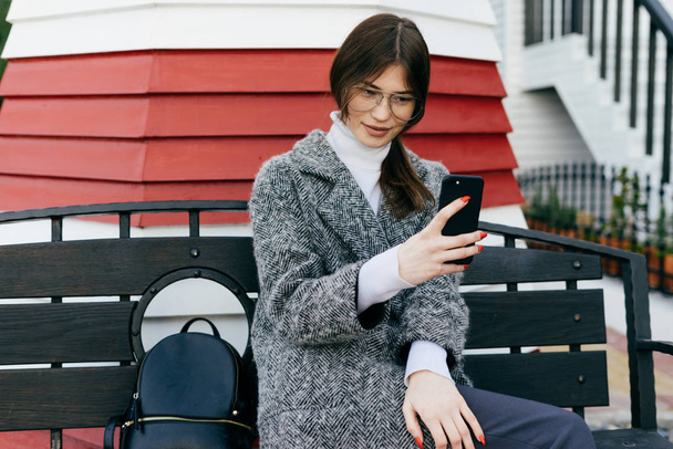 a stylish dark-haired girl with glasses and a gray coat sits on a bench in the open air and makes selfie - Photo, Image