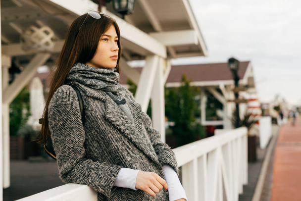 menina da cidade de cabelos escuros em um casaco cinza na moda desfrutando do tempo, andando ao ar livre, estilo de rua
 - Foto, Imagem