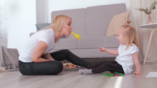 Attractive young blond mom inflate a balloon for her charming daughter sitting near the sofa. Family pastime. - Video