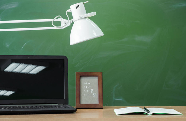 Teacher, student desk table. Education background. Education concept. Laptop with blank screen, copybook, pen, lamp and photo frame on the table with blackboard background. - Photo, image