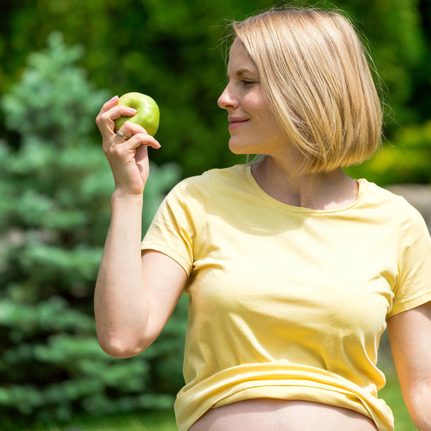 Mujer embarazada sentada en la hierba y sosteniendo una manzana en la mano
 - Foto, Imagen