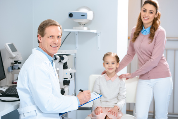 sonriente madre, hija y oftalmólogo mirando a la cámara en la sala de consulta oculista
 - Foto, imagen