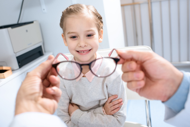 geschnittenes Bild eines Augenarztes, der lächelndem Kind in der Klinik eine neue Brille vorschlägt - Foto, Bild