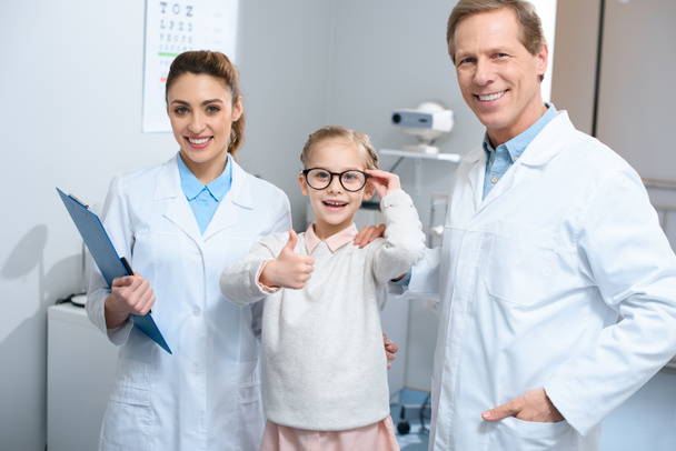 two smiling optometrists and little kid in eyeglasses showing thumb up - Foto, immagini