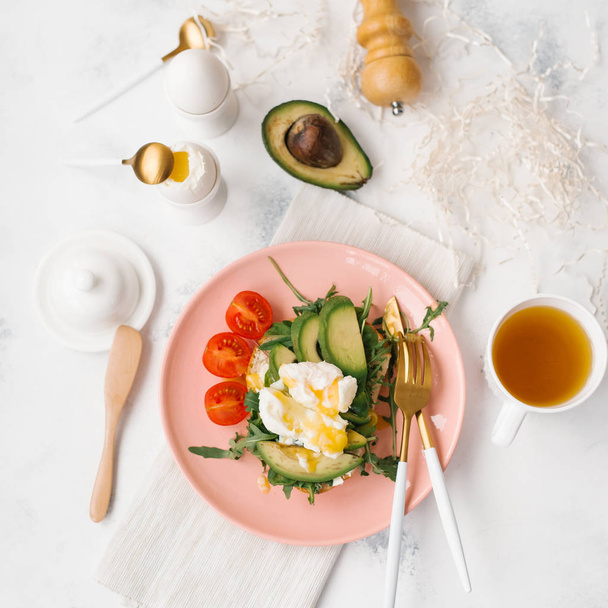 Food composition with sliced avocado, cherry tomatoes, arugula on pink plate with cutlery, eggs and tea cup - Photo, Image