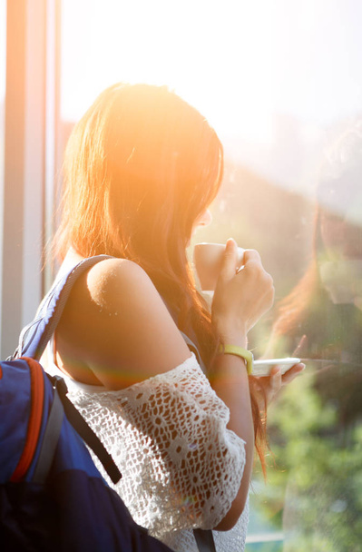Image of long-haired pensive woman with cup of tea and backpack by window - Foto, imagen
