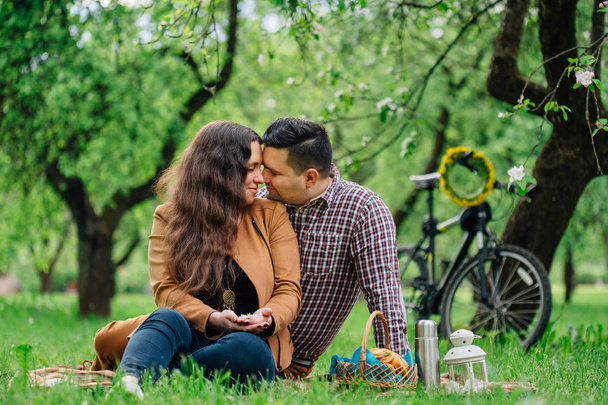 Young stylish happy couple in love having a picnic in blooming garden and bicycle behind. Pretty girl and man dating outdoors - Photo, image