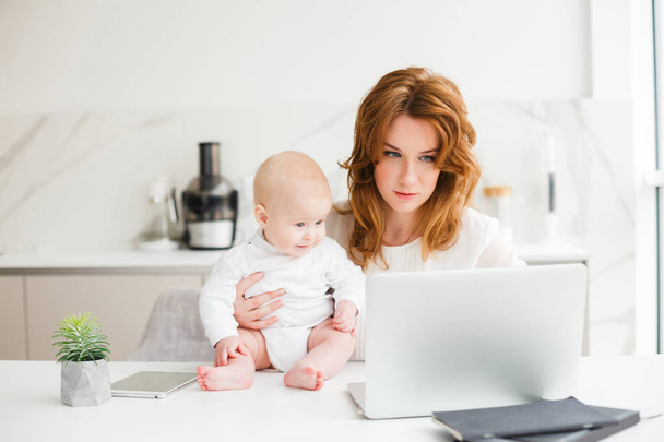 junge schöne Geschäftsfrau sitzt am Tisch und arbeitet am Laptop, während sie ihr süßes kleines Baby isoliert hält - Foto, Bild