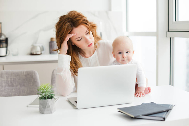 Porträt einer hübschen Geschäftsfrau, die am Tisch sitzt und am Laptop arbeitet, während sie ihr süßes kleines Baby isoliert hält - Foto, Bild