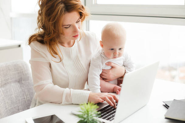 Portret Damy biznes siedział przy stole i pracę na laptopie trzymając swoje małe dziecko na białym tle - Zdjęcie, obraz