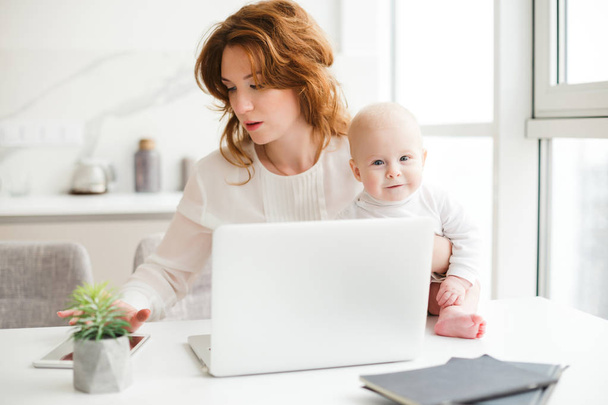 Портрет красивой женщины, сидящей за столом со своим маленьким ребенком и работающей с ноутбуком и планшетом изолированы
 - Фото, изображение