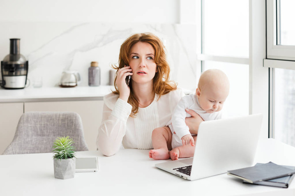 Portret przemyślany biznes kobieta siedzi przy stole z laptopa i mówić na jej telefon w ręku trzymając swoje małe dziecko na białym tle - Zdjęcie, obraz